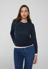 Suéter de punto azul marino con cuello redondo y mangas largas, con un dobladillo ajustado. Lleva una camisa blanca debajo y se combina con jeans azul oscuro.