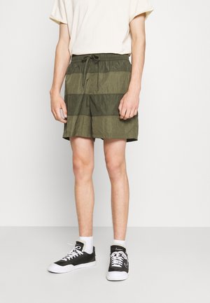 Personne portant un short rayé vert olive, un t-shirt blanc, des chaussettes blanches et des baskets noires et blanches, debout devant un fond blanc.