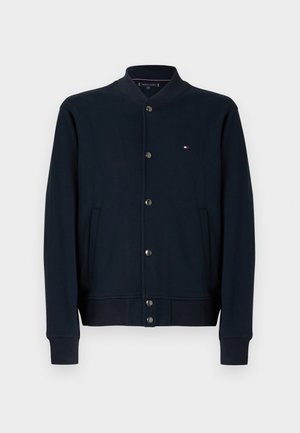 Tumesinine kapuutsi- või dressipluus, ees nööbitav, soonikkoes mansetid ja alumine äär, küljetaskud ning väike logo rinnal. Sile kangapind.