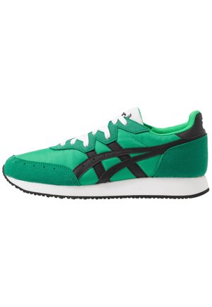 Baskets de sport vertes avec des rayures noires, des lacets blancs et une semelle intermédiaire blanche, présentées de profil sur fond blanc.