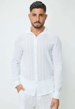 Homme aux cheveux courts et foncés et à la barbe, portant une chemise blanche texturée à boutons et un pantalon assorti, debout devant un fond uni.