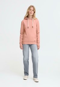 Sweatshirt à capuche rose avec cordon de serrage, texture douce, poignets côtelés, associé à un jean bleu clair et des baskets blanches.