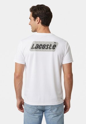 Hombre de espaldas que lleva una camiseta blanca de Lacoste con un gran logo y texto en la parte superior de la espalda y pantalones vaqueros azul claro.