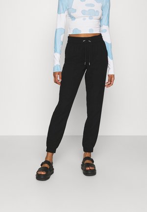 Kobieta w czarnych joggerach z elastycznymi mankietami, krótkim topie z niebiesko-białym wzorem chmur oraz czarnych sandałach z odkrytymi palcami.