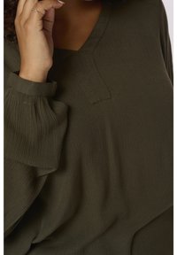 Blouse vert olive avec un col en V, en tissu texturé et manches bouffantes. Présente un design minimaliste et des détails de couture subtils.