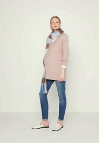Suéter rosa de malha oversized com decote em V, combinado com um cachecol azul claro com riscas castanhas e brancas, jeans azuis e sapatos brancos de slip-on.
