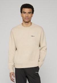 Beiger Rundhalspullover aus weichem Stoff, mit gerippten Bündchen und Saum sowie einem kleinen aufgestickten Logo auf der Brust.