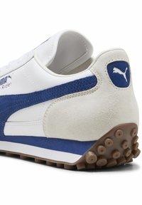 Baskets Puma avec une tige en cuir blanc, un accent en daim bleu, un talon beige et une semelle en caoutchouc avec des crampons ronds.