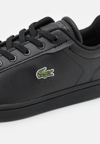 Baskets en cuir noir avec une surface texturée, un bout rond, des lacets noirs et un logo de crocodile vert brodé sur le côté.
