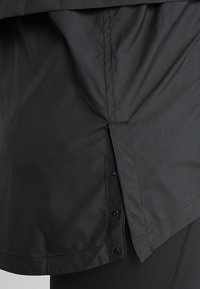 Chaqueta atlética negra con un diseño en capas, tela texturizada, detalles de botones a presión y un dobladillo trasero ligeramente alargado.