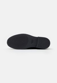 Semelle de chaussure noire avec un motif de grip texturé, fabriquée en caoutchouc avec un bord légèrement surélevé. Logo de la marque visible sur le côté.