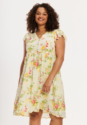 Lockenhaarige Frau, die lächelt und ein hellgelbes Blumenkleid mit flatternden Ärmeln und gesäumtem Saum trägt, steht vor einem schlichten Hintergrund.