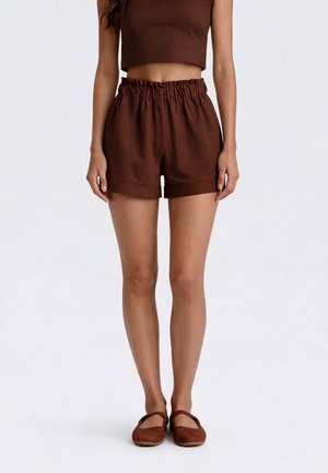 Donna con top corto marrone senza maniche, pantaloncini marroni a vita alta con fascia elastica in vita e scarpe basse marroni abbinate, in piedi dritta.