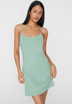 Femme aux longs cheveux noirs porte une robe sans manches vert clair avec de fines bretelles et un petit nœud au niveau de l'encolure, debout devant un fond uni.