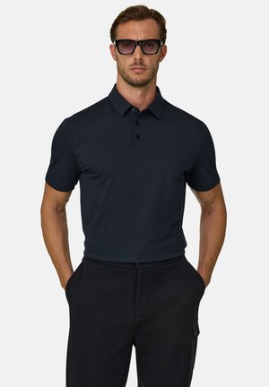 Mand iført en mørk navy kortærmet polo shirt, sorte bukser og mørke solbriller, står med hænderne i lommerne mod en hvid baggrund.