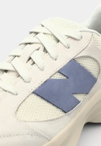 Sneaker med ljusgrå mockaöverkonstruktion, meshdetaljer, blå gummilogga och krämfärgade snören; har en dämpad sula och rund tåkonstruktion.