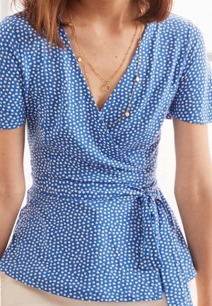 Haut enveloppant bleu avec des pois blancs, manches courtes, un lien à la taille et un décolleté en V. Texture lisse, tissu léger.