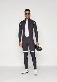 Casaco e calças de ciclismo pretos com detalhes brancos, combinados com uma camisola branca e ténis. Segurando um capacete preto com um design elegante.