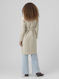 Casaco bege em mistura de lã com cintura cingida por um cinto, mangas compridas e corte direito, combinado com jeans azuis claros com folho e sapatos escuros.