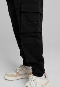 Svarta cargobyxor med flera fickor, med dragkedja. Bärs tillsammans med vita och beiga sneakers, som visar elastiska muddarna och materialets textur.