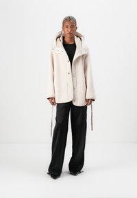 Beige fleece jacket with a hood, featuring snap buttons and drawstring accents, paired with black wide-leg pants. Smooth texture.