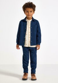 Jonge jongen staat, gekleed in een marineblauwe jas en broek, beige shirt met dierenprints, en bruine veterschoenen, tegen een effen witte achtergrond.