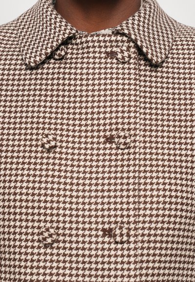Tuvums tuvtu attēls ar brūnu un baltu houndstooth rakstu divrindu mēteļa, ar atbilstošām auduma pogām un platu apkakli.