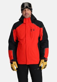 Rote und schwarze wasserdichte Jacke mit einer Reißverschlusstasche, gelben Handschuhen und grauen Akzenten. Entworfen für Aktivitäten bei kaltem Wetter.