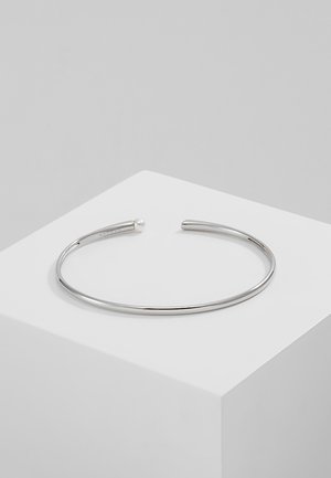 Bracelet manchette fin en argent avec un petit détail en perle à une extrémité, posé sur une surface blanche inclinée sur un fond gris.