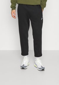 Person som bär svarta Nike-mjukisbyxor, vita sneakers med grå och neonkonturer samt en olivgrön sweatshirt, stående mot en enfärgad bakgrund.