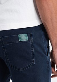 Jeans de mezclilla azul oscuro con un bolsillo trasero rectangular que presenta una etiqueta verde grabada con "Ombre Denim". Textura suave, costuras visibles.