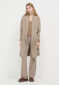 Frau mit langen blonden Haaren, die eine beige offene Strickjacke, ein braunes Knopfoberteil, weite beigen Hosen und dunkelbraunen Slipper trägt.