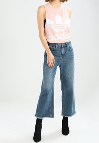 Kvinna iklädd ärmlös rosa Adidas-top, vida jeans som når till anklarna i blått, och svarta ankelstövlar, stående mot en enfärgad bakgrund.