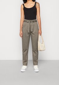 Hnědé a béžové kostkované kalhoty s uvázaným pásem, v kombinaci s černým topem na ramínka a bílými teniskami, držící krémovou kabelku přes rameno.