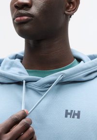 Ανοιχτό μπλε φούτερ με μικρό γκρι λογότυπο, διαθέτει κουκούλα με κορδόνι και υφή υφάσματος. Φορεμένο πάνω από πράσινο πουκάμισο.