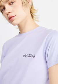 T-shirt in cotone lavanda con colletto rotondo, maniche corte e un logo ricamato nero sul petto, texture liscia e taglio casual.