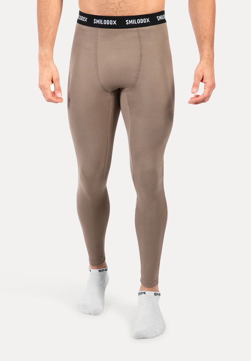Man gekleed in taupe compressieleggings met een zwarte tailleband met het label "SMILODOX" en witte enkelsokken met zwarte tekst.