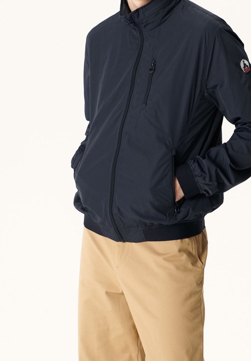 Veste légère bleu marine avec fermeture éclair avant, col montant, poches latérales et poignets élastiques, associée à un pantalon kaki. Texture lisse.