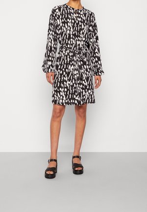 Vestido negro texturizado con un patrón abstracto blanco, mangas largas, escote en forma de ojal y cinturón atado en la cintura. Combinado con sandalias negras de plataforma.
