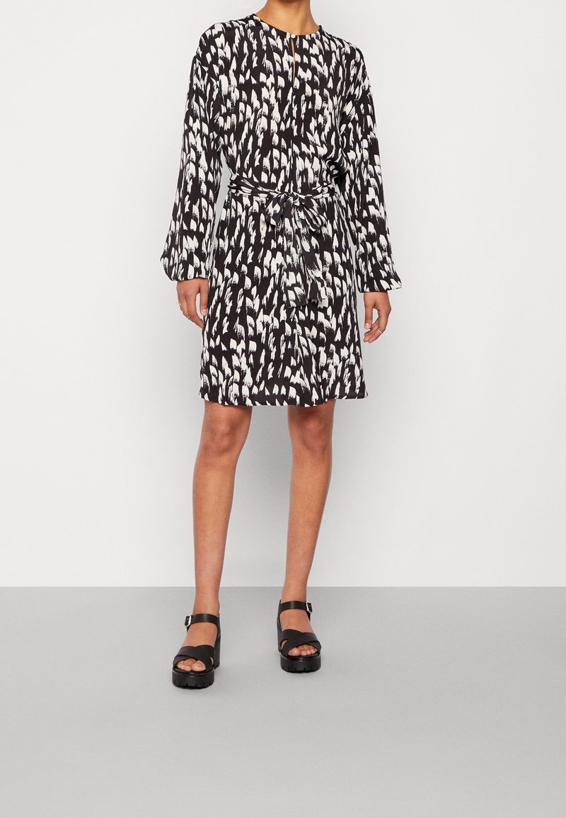 Czarna sukienka o fakturze z białym abstrakcyjnym wzorem, długimi rękawami, dekoltem w kształcie łezki i przewiązaną w talii paskiem. Noszona z masywnymi czarnymi sandałami.