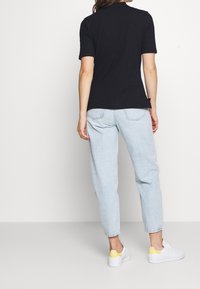 Femme portant un t-shirt noir à manches courtes, un jean taille haute bleu clair et des baskets blanches avec des accents jaunes au talon, se tenant de dos.
