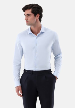 Camicia azzurra a maniche lunghe in tessuto liscio con colletto a punta e chiusura con bottoni, abbinata a pantaloni scuri su misura.