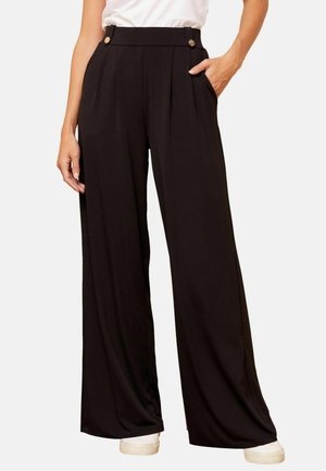 Frau trägt schwarze High-Waist-Hosen mit weitem Bein, goldenen Knöpfen und Seitentaschen, kombiniert mit weißen Sneakers und einem weißen Oberteil.