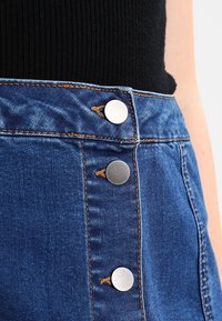 Gonna di jeans alta vita di colore blu con bottoni argentati e cuciture arancioni a contrasto lungo le cuciture. Tessuto liscio, design aderente.
