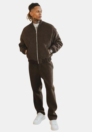 Jeune homme aux dreadlocks portant une veste marron zippée et un pantalon assorti, des baskets blanches et une montre, regardant sur le côté avec les mains dans les poches.