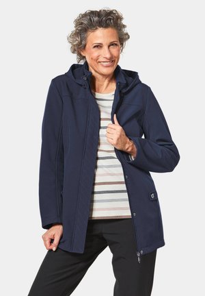 Navy softshell jacket with a hood, featuring quilted patterns and snap closures. Paired with a striped shirt and black pants.