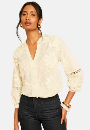 Femme portant un chemisier crème avec une grande broderie florale et des manches bouffantes, associé à un jean foncé taille haute et des boucles d’oreilles créoles dorées.