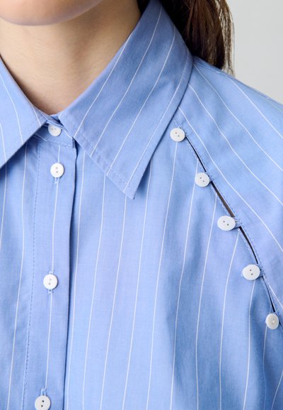 Camisa de botón azul claro con rayas blancas. Presenta un cuello con puntas y múltiples botones blancos en la tapeta delantera y en la manga.