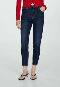 Jeans in denim blu scuro con vestibilità slim, vita media e design classico a cinque tasche. Indossati con una maglietta rossa aderente e sandali neri.