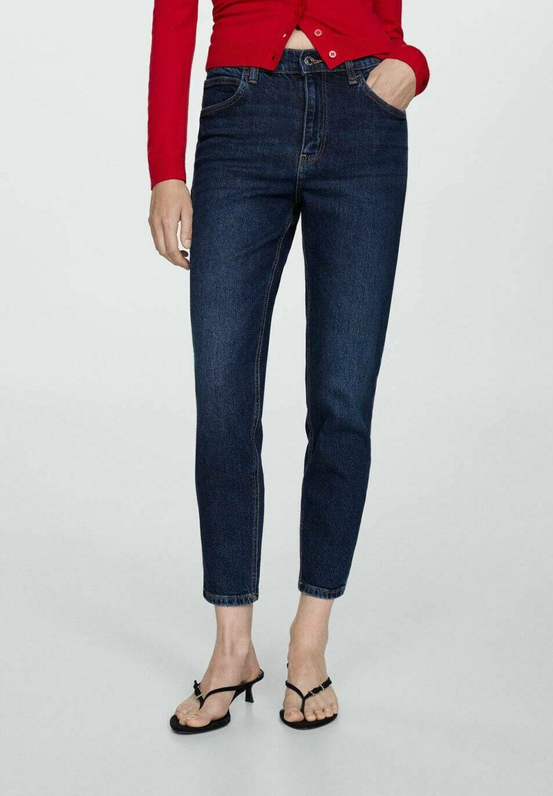 Jeans in denim blu scuro con vestibilità slim, vita media e design classico a cinque tasche. Indossati con una maglietta rossa aderente e sandali neri.